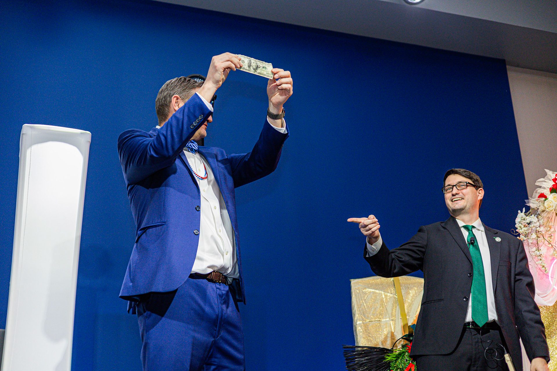A blindfolded volunteer holds a hundred dollar bill above his head while Josh Weidner points, on stage at the GiGi's Playhouse Indy gala.