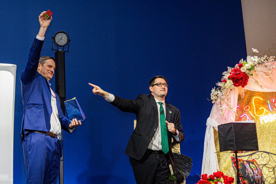 Josh Weidner pointing towards a volunteer in a blue suit who is holding a solved Rubik's cube above his head during the stage set at the GiGi's Playhouse Indy gala.
