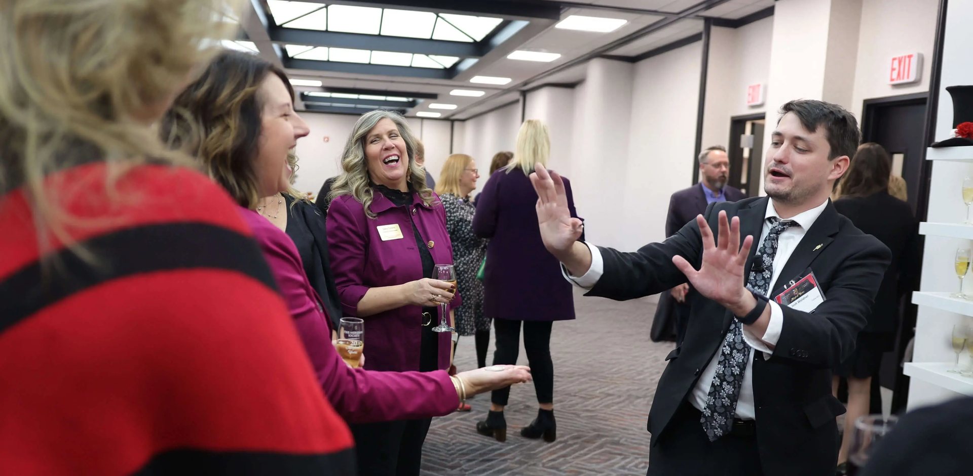 Josh Weidner performing close-up magic for guests holding drinks at The Meridian Society twentieth anniversary reception, with the champagne display behind them.