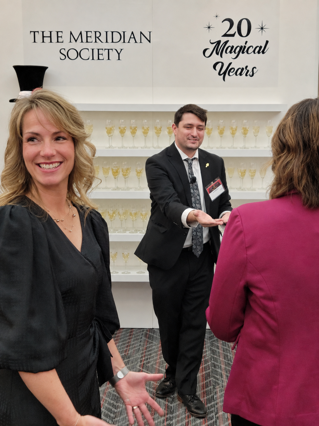 A guest smiling towards the camera while Josh Weidner performs close-up magic behind her at The Meridian Society twentieth anniversary reception.